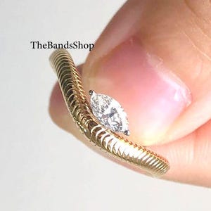 May include: A gold ring with a marquise-cut diamond. The ring band has a textured, ridged design. The ring is held up against a white background. The text "TheBandsShop" is visible.