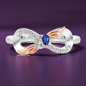 May include: A silver ring with a unique infinity design, featuring a blue pear-shaped gemstone and a clear, round-cut stone. The ring is accented with small, sparkling crystals and rose gold-colored wings. TheBandShop logo is visible.