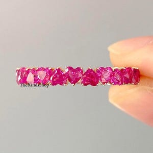 May include: A gold ring featuring a series of heart-shaped, vibrant pink gemstones. The gemstones are set closely together, creating a continuous band of color. The ring is held against a soft gray background.