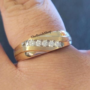 Puede incluir: Un anillo dorado con una fila de seis piedras claras. El anillo tiene un diseño en capas y un acabado pulido. El texto "TheBandsShop" es visible en el anillo.