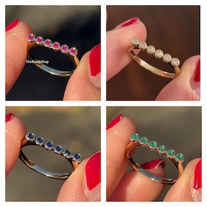 Peut inclure: Une bague en or ornée d'une rangée de cinq pierres précieuses rondes serties clos. L'image présente quatre variantes de la bague avec des pierres de couleurs différentes : rose vif, blanc transparent, bleu profond et vert éclatant. Le texte TheBandsShop est visible.