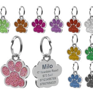 May include: A selection of silver paw print dog tags with glitter accents in various colours, including green, orange, pink, purple, red, silver, gold, blue, and black. One tag features the name "Milo" and contact details.