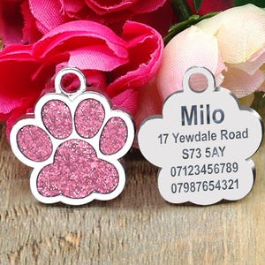 May include: Two silver dog tags, one shaped like a paw print with pink glitter and the other a cloud shape. The paw print tag is blank. The cloud tag has the name "Milo", address "17 Yewdale Road", postcode "S73 5AY", and phone numbers "07123456789" and "07987654321".