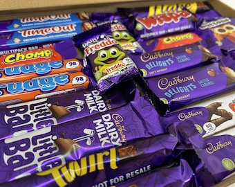 May include: An assortment of chocolate bars in various wrappers, including Cadbury Dairy Milk, Twirl, Freddo, and Cadbury Delights. The bars are arranged in a box and feature a variety of colors and designs.
