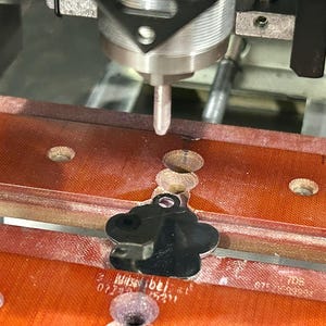 May include: A close-up of a CNC machine engraving a silver paw print pet tag. The tag is being held in place by a red and brown clamp.