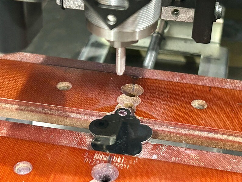 May include: A close-up of a CNC machine engraving a silver paw print pet tag. The tag is being held in place by a red and brown clamp.