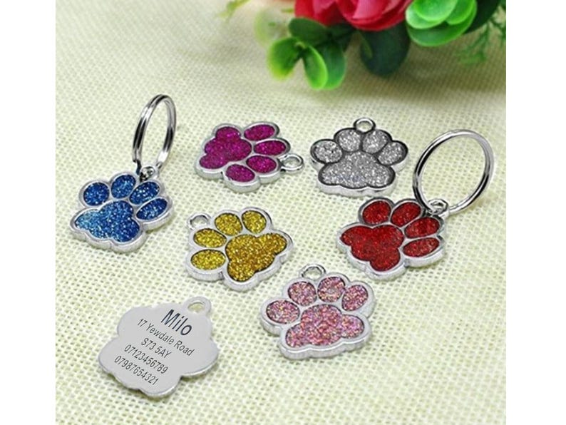 May include: Six silver paw print dog tags with glitter accents in blue, red, pink, gold, silver, and purple. The tags are attached to key rings. One tag has a silver engraved tag with the text "Milo, 17 Yewdale Road, ST3 5AY, 07121456789, 07981654321".