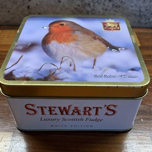 May include: A rectangular tin with a gold rim and a lid featuring a Red Robin bird in a snowy scene. The tin's side displays "STEWART'S Luxury Scottish Fudge WHITE EDITION" in red and white text.