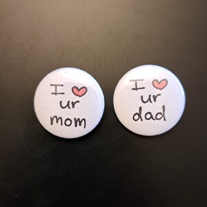 May include: Two white button pins with black text and a red heart. The text on the left pin reads "I ♥ ur mom" and the text on the right pin reads "I ♥ ur dad".