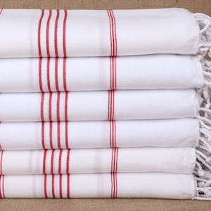 May include: A stack of six white towels with red stripes and white fringe. The towels are folded and stacked on a brown surface.