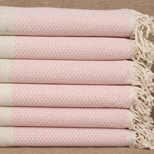 Puede incluir: Una pila de toallas rayadas de color rosa y blanco con flecos en los bordes. Las toallas están dobladas cuidadosamente y tienen un sutil patrón rosa.