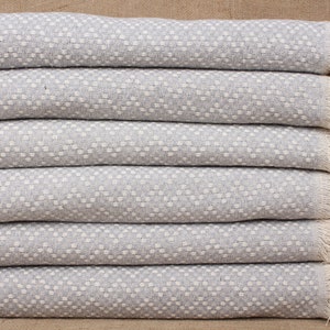 May include: A stack of six pale grey blankets with a textured, dotted pattern. Each blanket has a fringe along one edge. The blankets are neatly folded and stacked on top of each other.