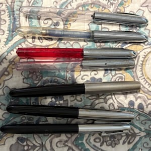 May include: A collection of seven pens arranged horizontally. The set includes two clear pens, one with a blue ink cartridge, one translucent red pen, one translucent pink pen, and three black pens with silver-toned caps and clips.
