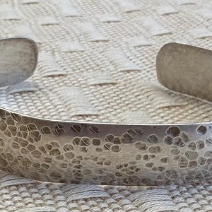 May include: Silver-toned cuff bracelet with a hammered texture. The open cuff design features rounded ends. The bracelet's surface has a dimpled pattern, creating a textured appearance. This jewelry piece is suitable for everyday wear.