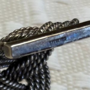 May include: Close-up of a silver-toned bracelet with a braided rope design. The rectangular clasp is marked "STERLING". The bracelet is on a white, textured surface.