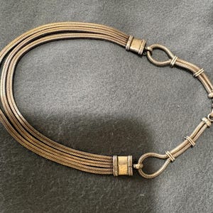 May include: A vintage silver-toned necklace with a multi-strand chain design. The necklace features a series of woven strands that connect to a decorative clasp. The clasp has a loop design and a hook closure. The necklace is displayed on a gray surface.