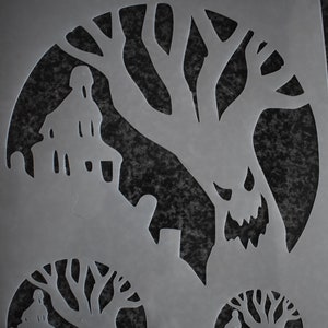 Può includere: Uno stencil di plastica grigia con il testo "GET STENCILLED" in alto. Lo stencil presenta tre disegni di una casa infestata da fantasmi con un albero e un viso di mostro.