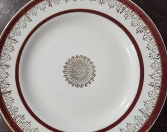 May include: White plate with a burgundy border and gold filigree detailing. The center of the plate features a gold circular design. The plate has a classic, elegant aesthetic.