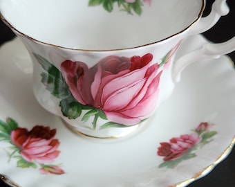 May include: A white teacup and saucer with a pink rose design. The cup has a gold rim and the saucer has a scalloped edge.