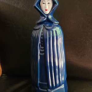 May include: A blue ceramic figurine of a woman wearing a hooded robe. The woman has a white face with painted black eyes and red lips.