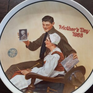 Puede incluir: Un plato de cerámica blanco con una ilustración colorida de una madre y un hijo. El hijo sostiene una foto de una niña. El texto "Mother's Day 1988" está impreso en rojo en el plato. El nombre del artista "Norman Rockwell" está impreso en marrón en la parte inferior del plato.