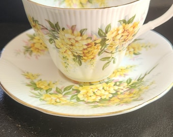 May include: A white teacup and saucer with a floral pattern featuring yellow and pink flowers. The cup and saucer have a gold trim.