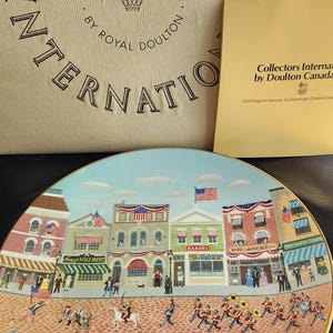 May include: A decorative plate from the Collectors International series by Royal Doulton. The plate features a colorful town scene with buildings, people, and an American flag. The plate is in a box with the words "Collectors International" and "Fine Art on Fine China."