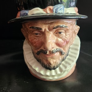 May include: A ceramic mug shaped like a man's head wearing a black hat with a white ruffle collar. The man has a brown beard and moustache.