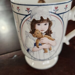 Könnte beinhalten: Weiße Keramiktasse mit blauem und rotem Rand. Die Tasse zeigt ein Mädchen, das einen Teddybär hält. Das Mädchen trägt ein weißes Kleid mit einem blauen Band und einer weißen Haube.
