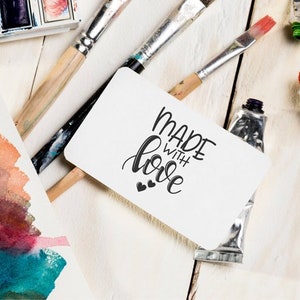 May include: A white card with the text "Made with love" in black calligraphy. The card is surrounded by paint tubes, paintbrushes, and watercolour paper.
