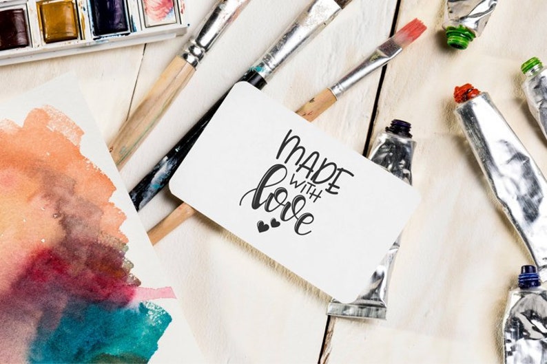 May include: A white card with the text "Made with love" in black calligraphy. The card is surrounded by paint tubes, paintbrushes, and watercolour paper.