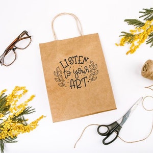 May include: A brown paper bag with a white background. The bag has a handle and the text "LISTEN to your ART." printed on it. There are sprigs of yellow mimosa flowers, a spool of twine, and a pair of scissors on the table.