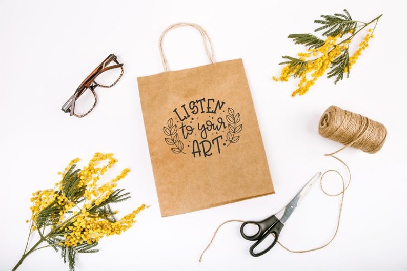 May include: A brown paper bag with a white background. The bag has a handle and the text "LISTEN to your ART." printed on it. There are sprigs of yellow mimosa flowers, a spool of twine, and a pair of scissors on the table.