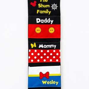 May include: A black fabric wall organizer with multiple pockets, featuring the text "The Shum Family." Each pocket is a different color and labeled with names like "Daddy," "Mommy," and "Wesley." Decorative elements include Mickey Mouse and bow tie graphics.