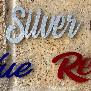 May include: Five metal words, "Black", "Silver", "Copper", "Blue", and "Red", are displayed on a stone wall. Each word is painted in a different color, matching the word.