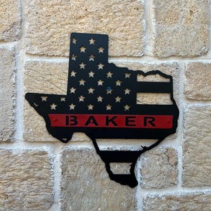 May include: Black metal cutout of the state of Texas with an American flag design and a red stripe across the bottom with the name "BAKER" in white letters.