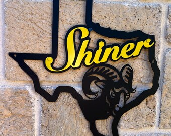 May include: Black metal cutout of the state of Texas with a gold "Shiner" logo and a black ram's head inside the state outline.