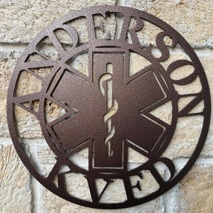 May include: A brown metal sign with a Star of Life symbol in the center. The words "Dedicated" and "Served" are written around the symbol.