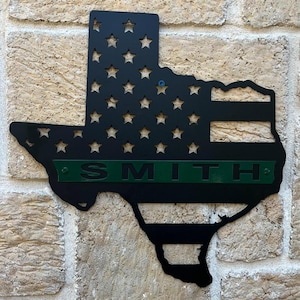 May include: Black metal cutout of the state of Texas with the American flag design and a green stripe across the middle with the name "SMITH" in white letters.