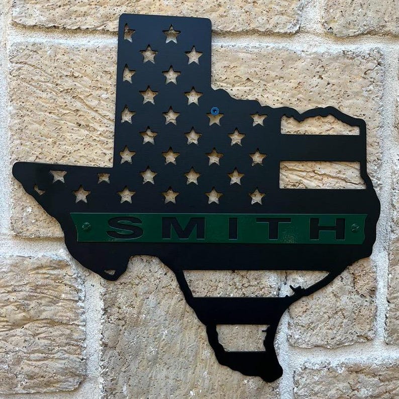May include: Black metal cutout of the state of Texas with the American flag design and a green stripe across the middle with the name "SMITH" in white letters.