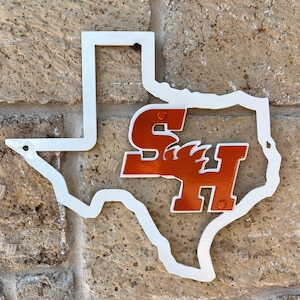 May include: A white metal cutout of the state of Texas with an orange and white logo of the letters "SH" inside. The metal sign is mounted on a brick wall.