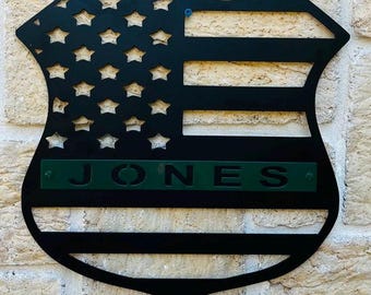 May include: Black metal shield-shaped sign with a cutout of the American flag and the name "JONES" in green lettering.