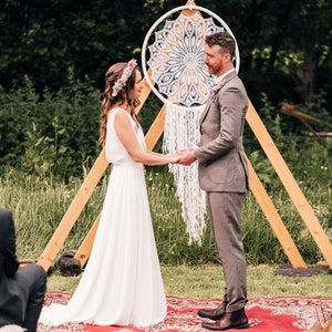 May include: An outdoor wedding ceremony featuring a bride in a white dress and a groom in a grey suit. They stand on a red rug, holding hands, with a decorative dreamcatcher. Guests are seated on hay bales.