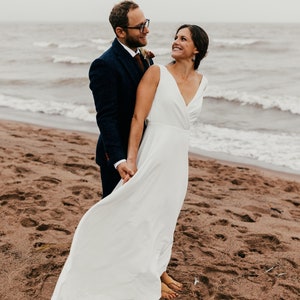May include: A couple on a sandy beach, the woman in a white wedding dress and the man in a navy blue suit. They are holding hands and looking at each other. The sea is in the background under a cloudy sky.