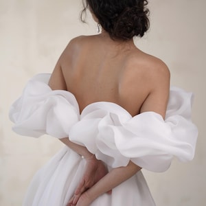 Könnte beinhalten: Ein weißes schulterfreies Brautkleid mit voluminösen, gerüschten Ärmeln. Das Kleid hat einen weiten Rock. Die Person, die das Kleid trägt, hat dunkles Haar, das zu einem Dutt gebunden ist. Der Hintergrund ist hellbeige.