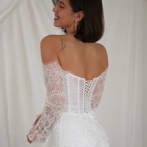 May include: A white, off-the-shoulder wedding dress with long, lace sleeves. The bodice features a corset-style back with delicate lacing. The dress is adorned with intricate lace detailing and subtle beading, creating a romantic and elegant look.
