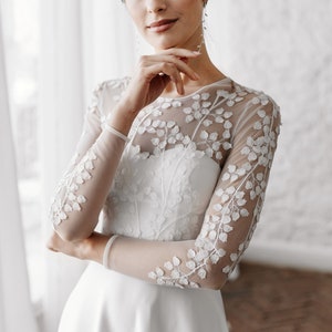 May include: A white wedding dress with long, sheer sleeves adorned with delicate floral embroidery. The dress features a fitted bodice and a flowing skirt, creating an elegant silhouette. The bride is wearing long, dangling earrings.