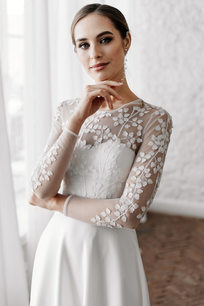 May include: A white wedding dress with long, sheer sleeves adorned with delicate floral embroidery. The dress features a fitted bodice and a flowing skirt, creating an elegant silhouette. The bride is wearing long, dangling earrings.