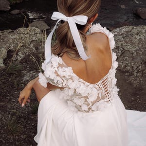 K&ouml;nnte beinhalten: Ein wei&szlig;es Hochzeitskleid mit floralen Spitzen&auml;rmeln und tiefem R&uuml;ckenausschnitt. Das Kleid hat einen R&uuml;cken im Korsett-Stil und wird mit einem wei&szlig;en Band im Haar kombiniert. Die Kulisse ist im Freien, in der N&auml;he eines felsigen Gebiets und eines Baches.