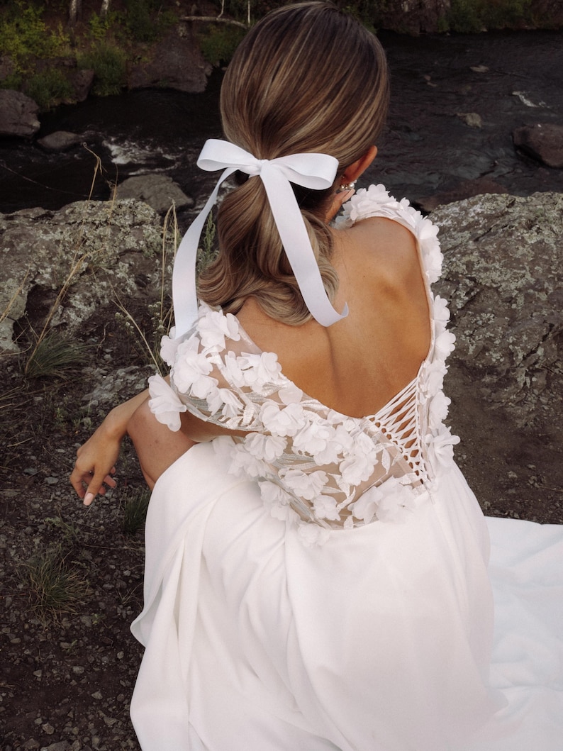 K&ouml;nnte beinhalten: Ein wei&szlig;es Hochzeitskleid mit floralen Spitzen&auml;rmeln und tiefem R&uuml;ckenausschnitt. Das Kleid hat einen R&uuml;cken im Korsett-Stil und wird mit einem wei&szlig;en Band im Haar kombiniert. Die Kulisse ist im Freien, in der N&auml;he eines felsigen Gebiets und eines Baches.